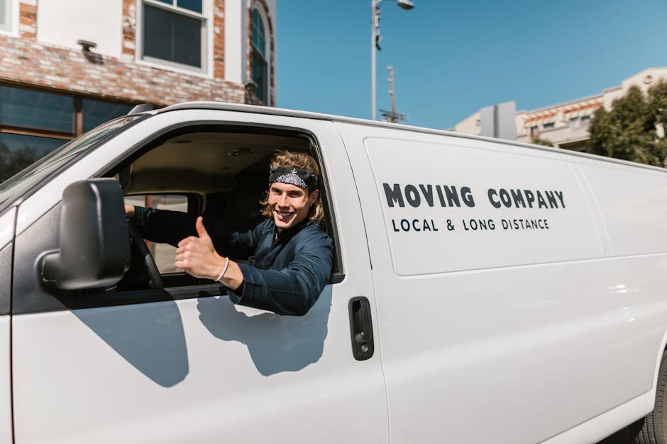 A young man with a beard, wearing a red cap backwards, a beige and brown jacket over a checked shirt, and fingerless gloves, sits at the rear of a van with its doors open. Inside the van, there are se