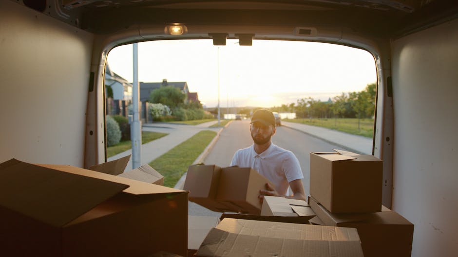 A professional mover from Man with Van Brompton is loading cardboard boxes and packing materials into the rear of a white panel van outside a modern building with large glass windows. The mover, weari