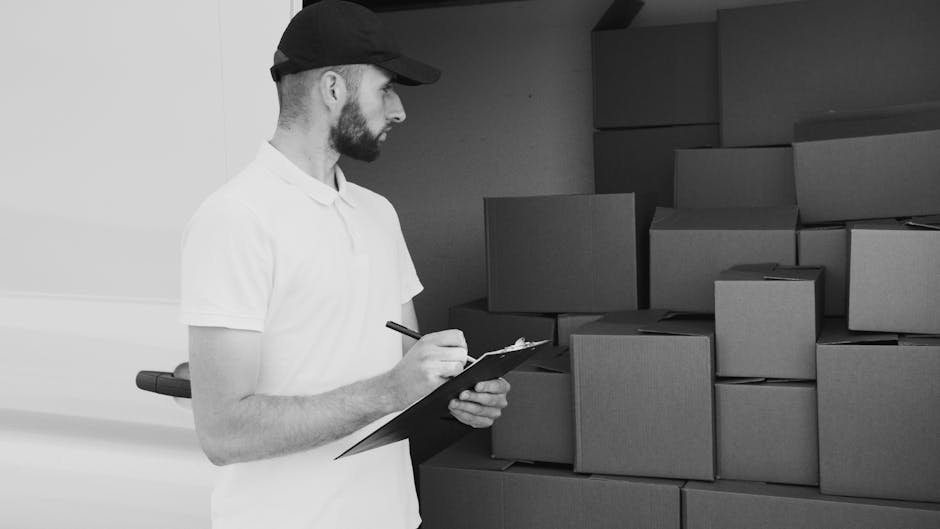 A man with a beard, wearing a white polo shirt and a black cap, is standing indoors next to a large stack of cardboard boxes during a home relocation process. He is holding a clipboard and pen, appearing to take inventory or check items before packing or loading. The boxes are of various sizes, neatly stacked against a plain wall, some with visible packing tape, and are likely filled with household items. The scene is well-lit, with natural light or interior lighting highlighting the workspace. This image relates to professional removals services, such as those provided by Man with Van Brompton, that specialise in packing, furniture transport, and home relocation logistics, capturing the preparatory phase of a house move.