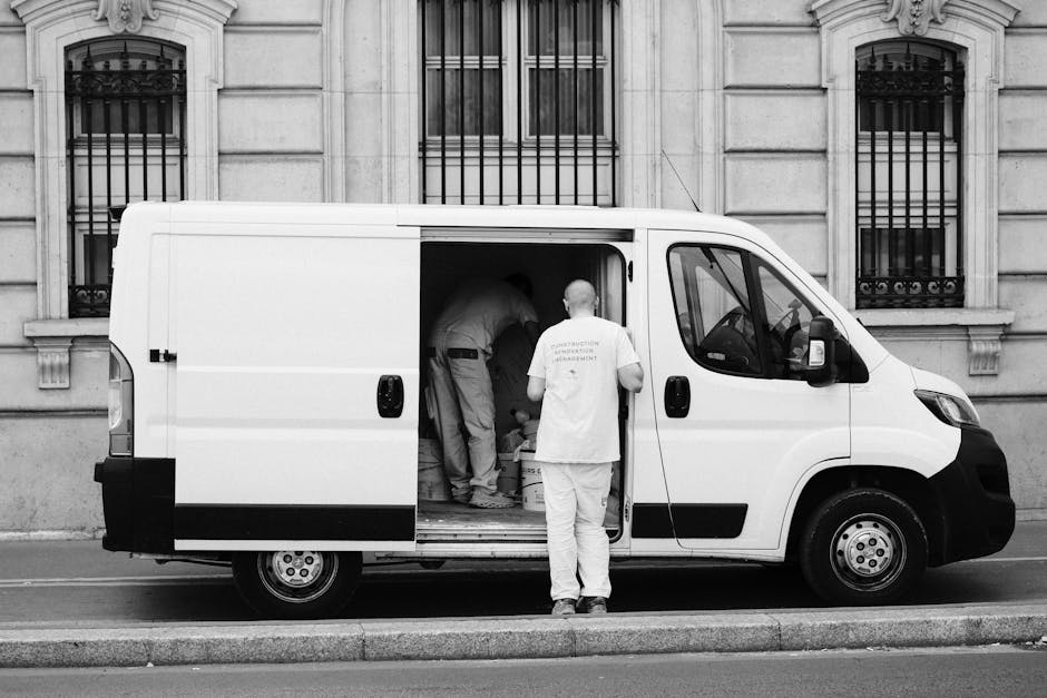 A black and white photograph showing a white delivery van parked on a street in front of a building with stone façade and barred windows. The side door of the van is open, revealing several workers inside and around the vehicle engaged in a home relocation or furniture transport process. One worker is standing outside the van, facing away, wearing a T-shirt with text on the back, while others are inside carrying and arranging packed boxes, some wrapped in plastic or placed in cardboard cartons. The van's interior contains additional boxes and packing materials, and the workers are using various equipment such as trolleys and straps to facilitate the loading process. The scene is set in an urban environment, possibly during a furniture move or packing and moving service by Man with Van Brompton, known for house removals and relocation services in the area.